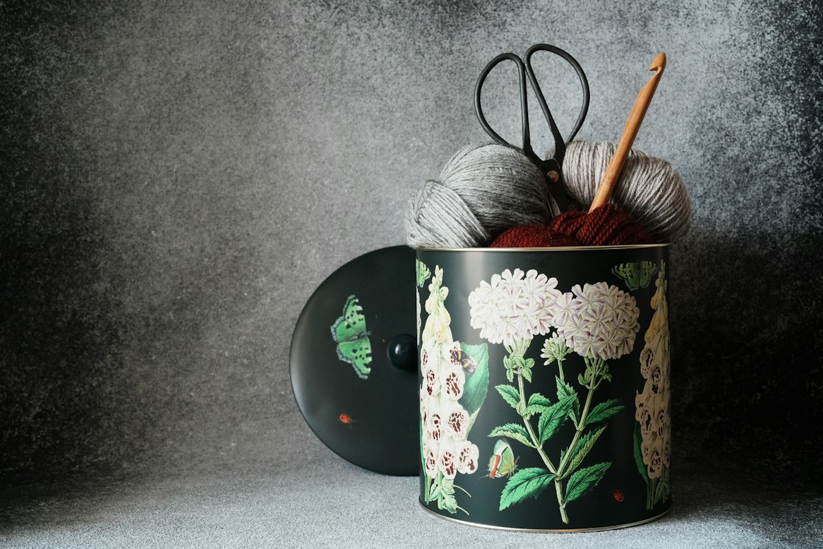Grey and burgundy yarn, scissors and a wooden crochet hook in a floral tin